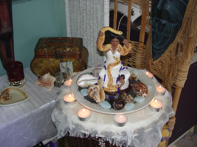A voodoo offering of lit candles around a sculpture of a woman with a snake on an altar
